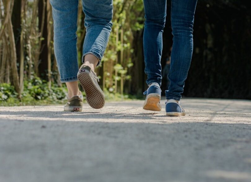 Två personer promenerar på en grusväg genom skogen i jeans och sneakers